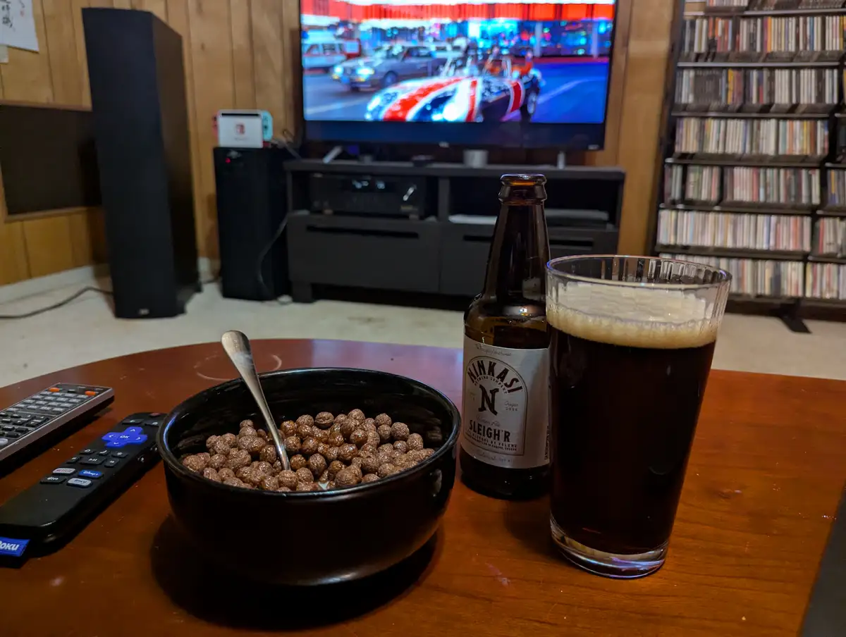 A cozy living room scene shows a wooden table with a bowl of chocolate cereal, a bottle of Ninkasi Sleigh'R Winter Ale, and…