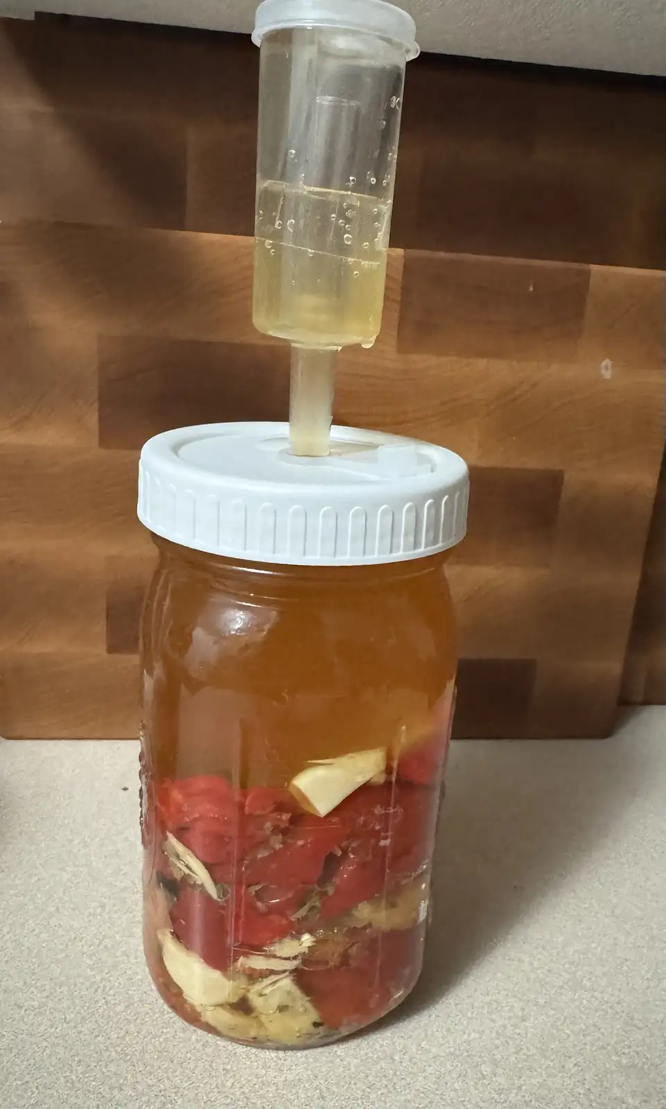 A glass mason jar filled with chopped red tomatoes and sliced garlic cloves submerged in liquid, sitting on a countertop. A…