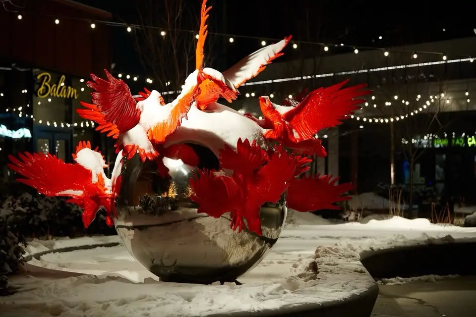 A nighttime photo of a red glass sculpture of multiple birds bursting from a reflective sphere, all covered in snow. The…