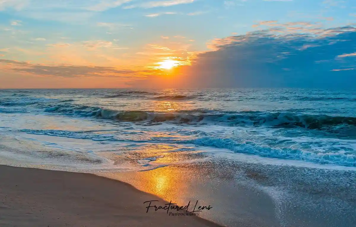 A serene beach sunset photo shows waves rolling onto sand under a sky painted with orange and blue hues, with a watermark…