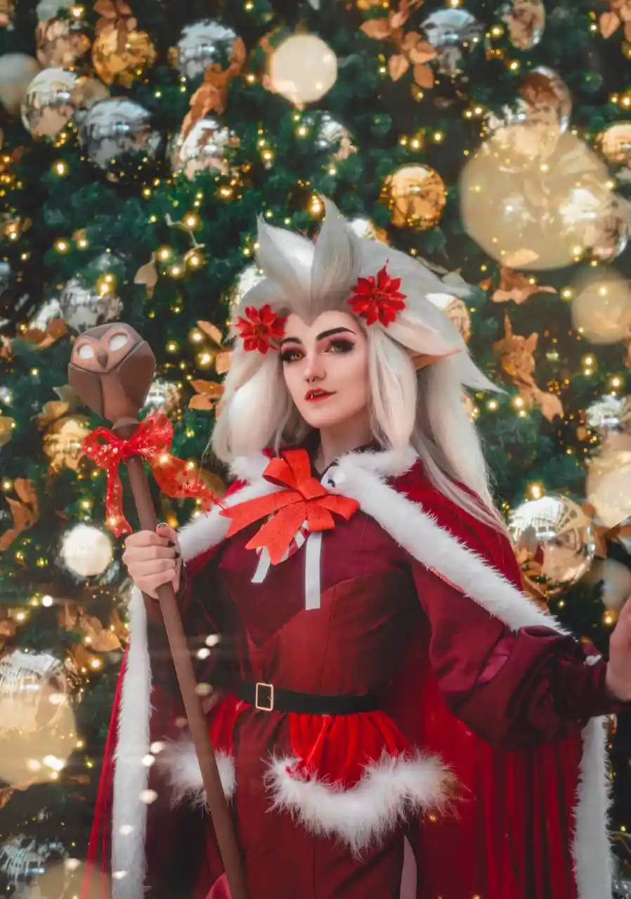 A person in a festive red and white costume with white hair, red flowers in their hair, and holding a staff with an owl…