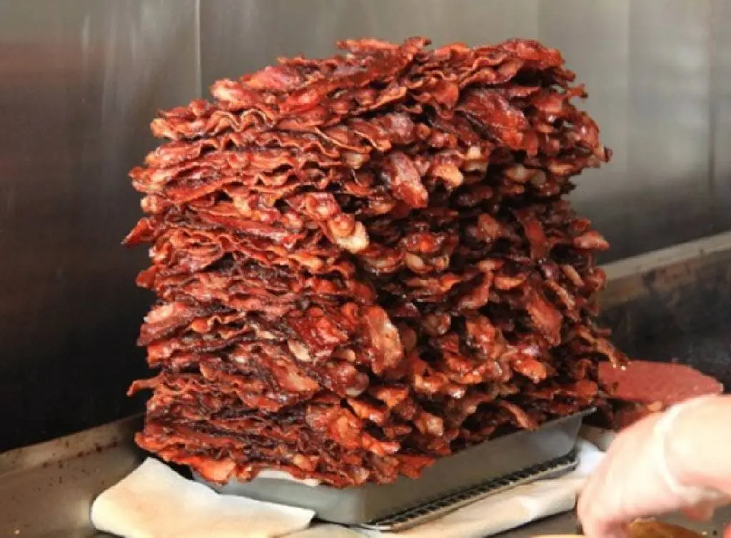 A towering, greasy pile of cooked bacon strips sits in a metal tray on a commercial kitchen counter, with a gloved hand…