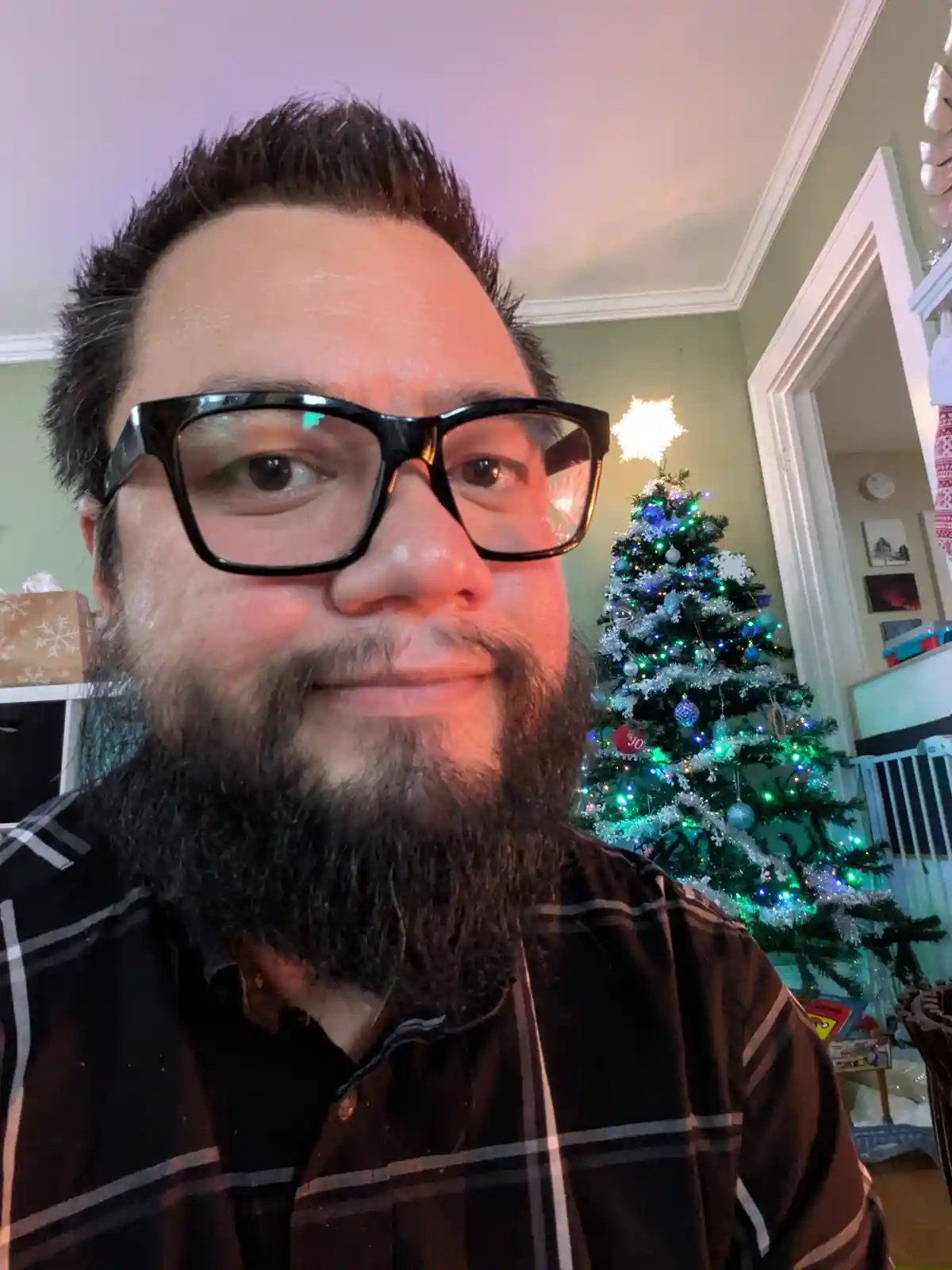 A man with a beard and black-rimmed glasses smiles at the camera while standing in front of a decorated Christmas tree with…
