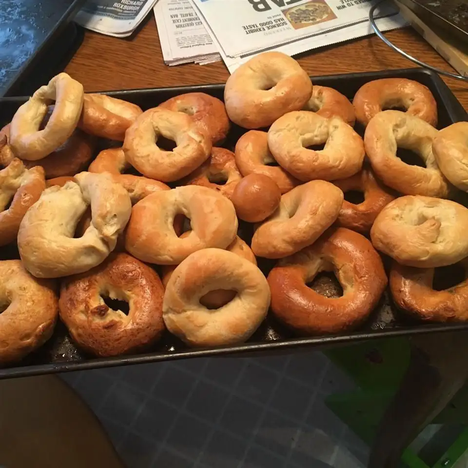 A black baking tray is filled with dozens of golden-brown, freshly baked bagels, some perfectly round and others misshapen…