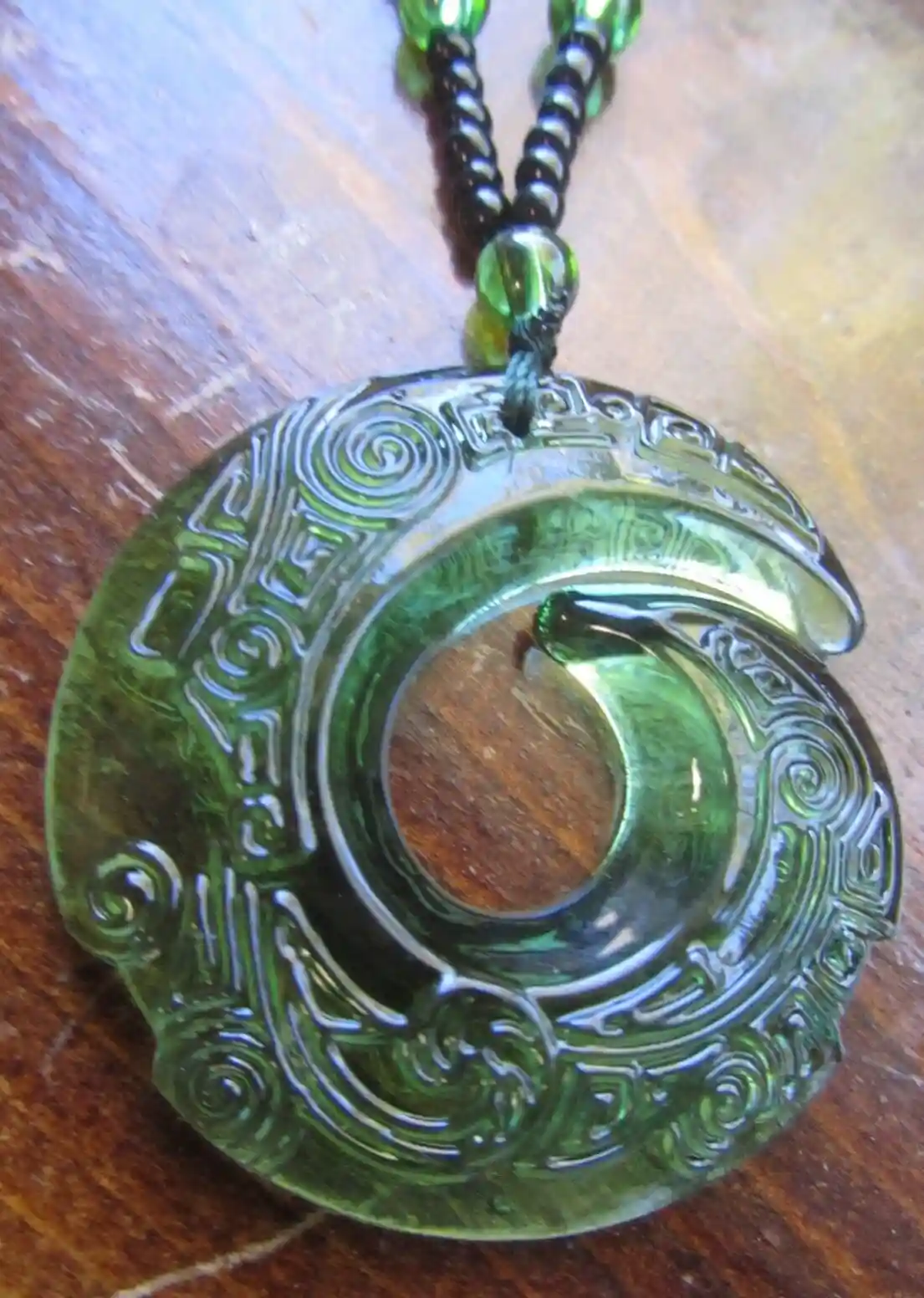 A close-up photo shows a green, carved, circular pendant with intricate spiral and geometric patterns, resembling a…
