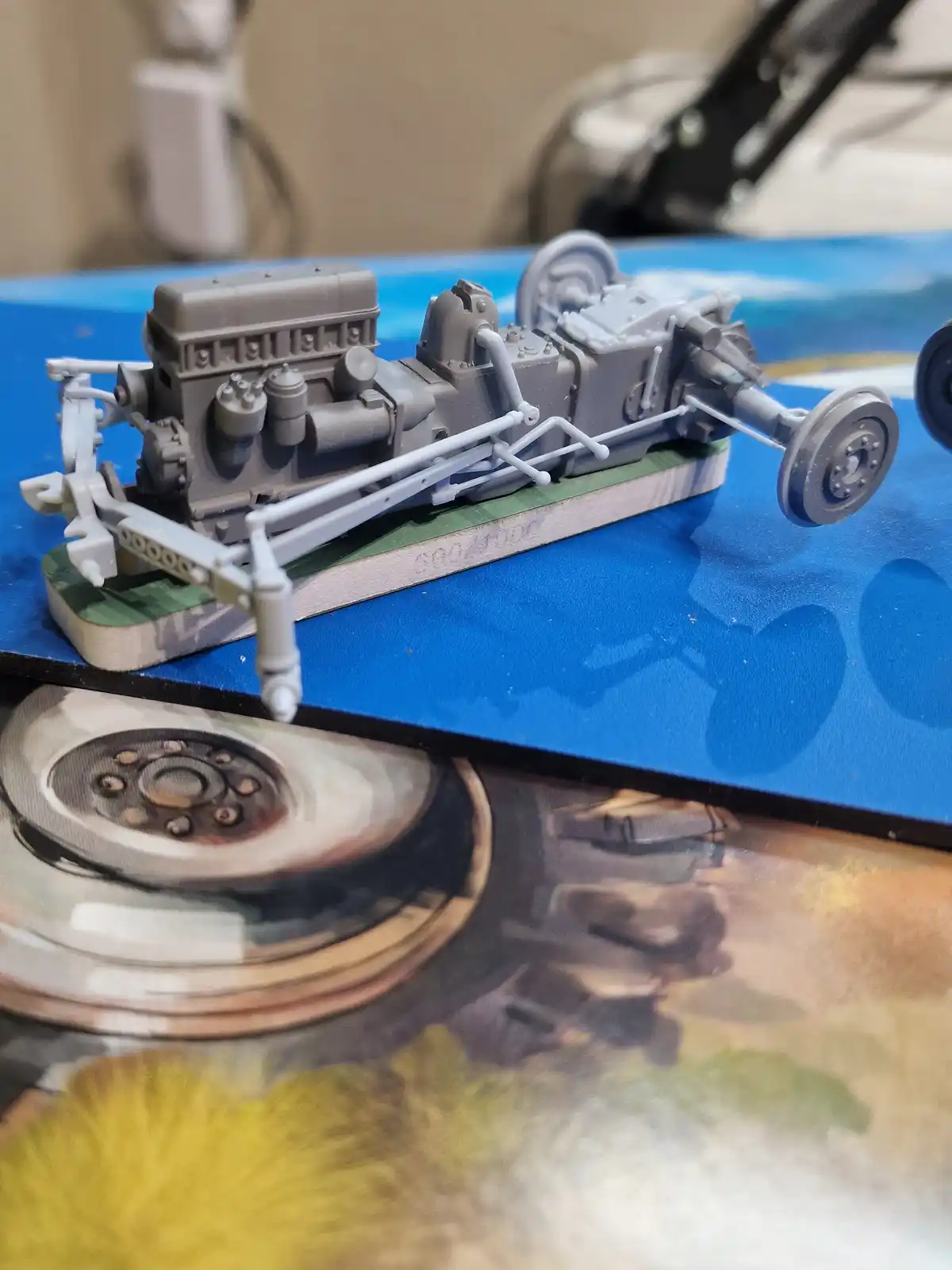 A gray, unassembled plastic model kit of a car's engine and chassis sits on a blue work surface, with a printed image of a…