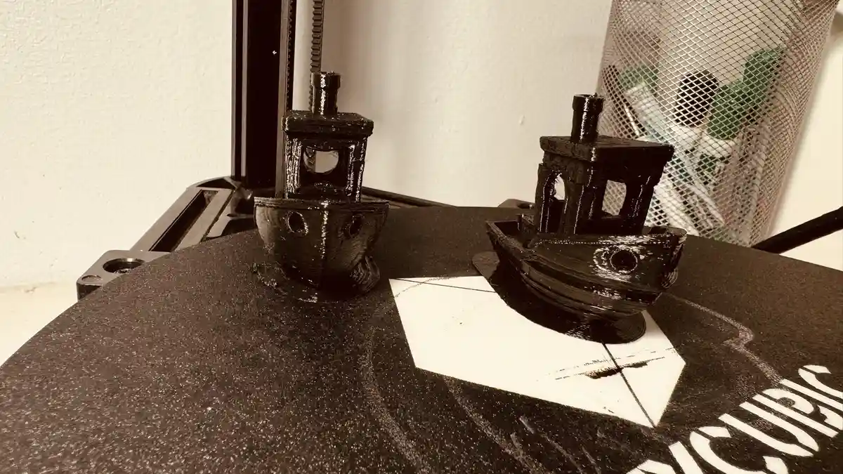 Two small, black 3D-printed boat models sit on a printer bed, appearing to face each other as if in conversation. The image…