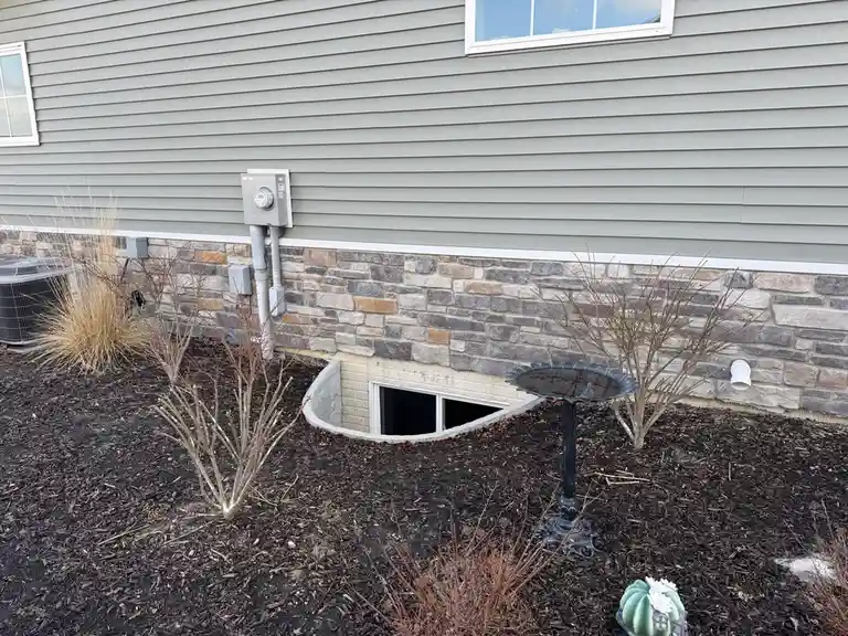 An exterior shot of a house with gray siding and a stone foundation, featuring a small, curved basement window set into the…