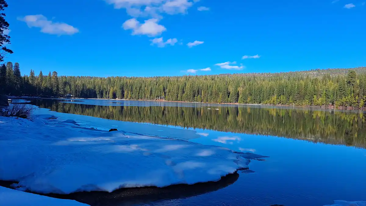 A serene winter landscape shows a partially frozen lake with snow-covered shorelines and a clear blue sky reflected in the…