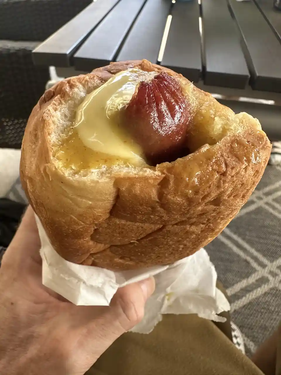 A hand holds a golden-brown bread roll with a glossy, reddish-brown sausage nestled inside, topped with a generous amount of…