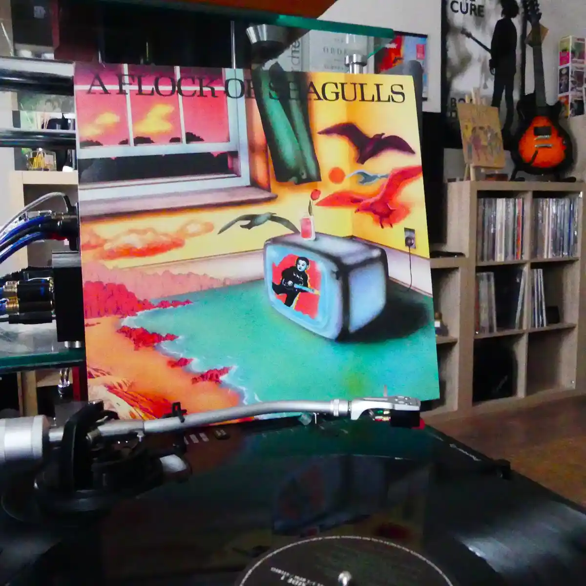 A vinyl record player with a needle arm is positioned in front of the album cover for 'A Flock of Seagulls,' which depicts a…