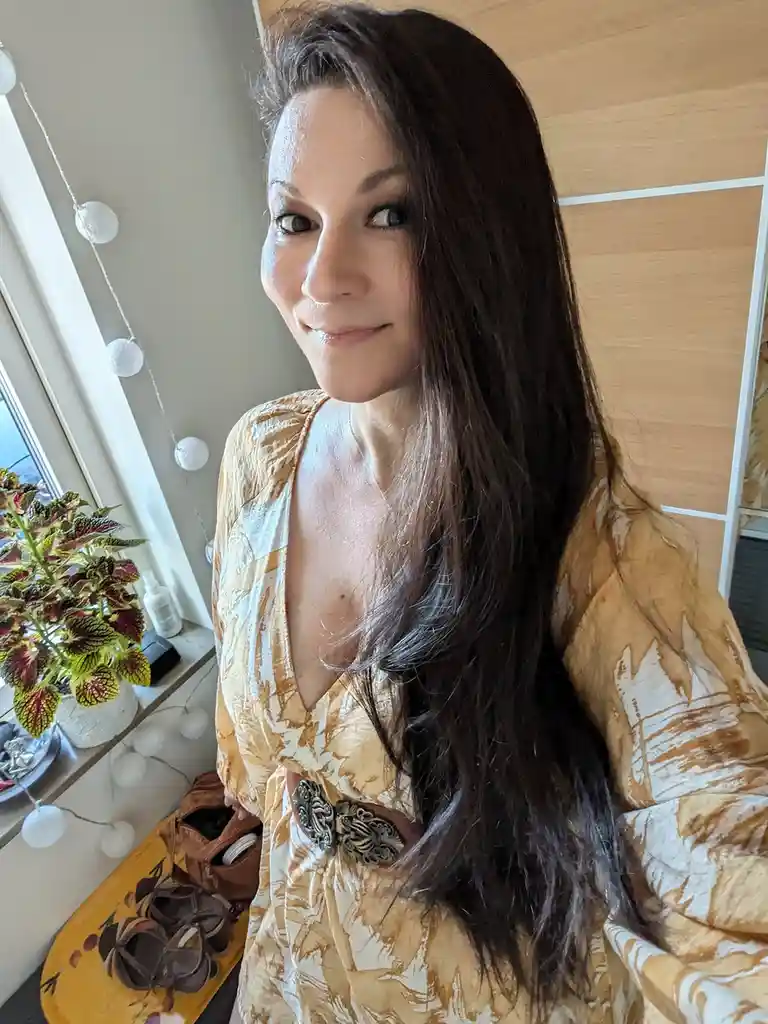 A woman with long dark hair wearing a patterned yellow and white dress with a decorative belt smiles at the camera while…