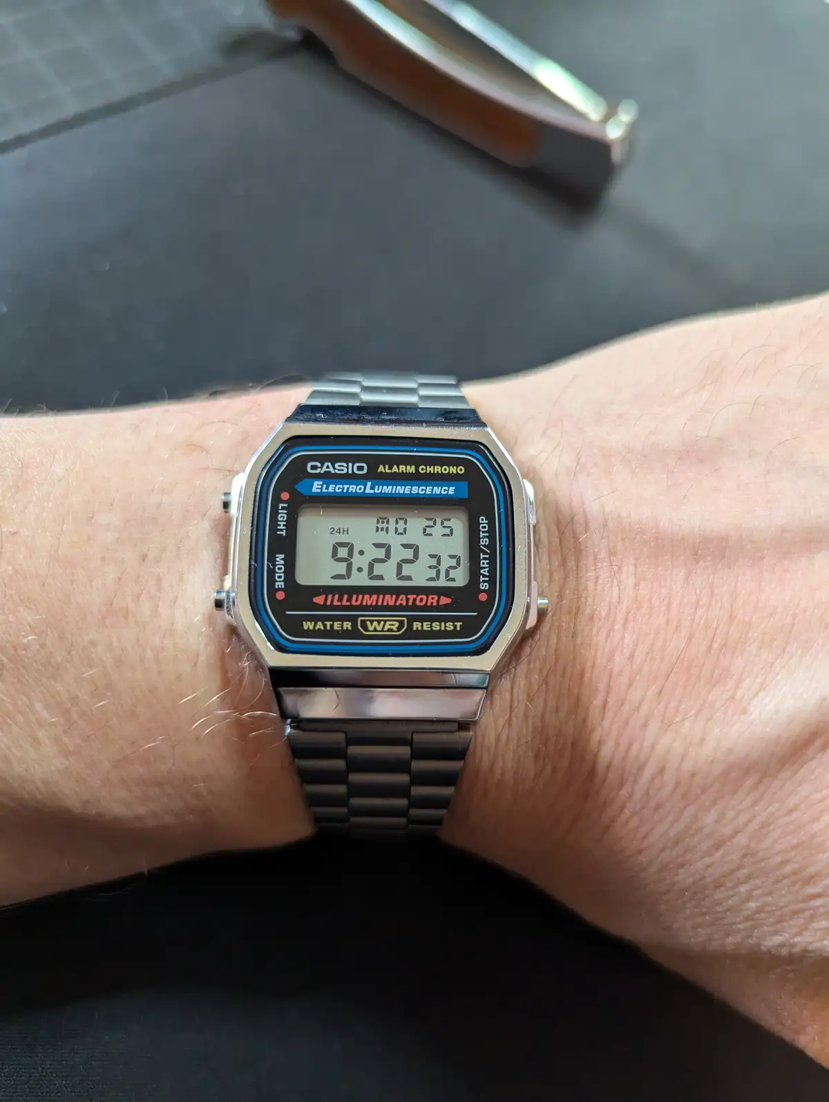 A close-up photo shows a person's wrist wearing a vintage Casio digital watch with a silver metal band. The watch face…