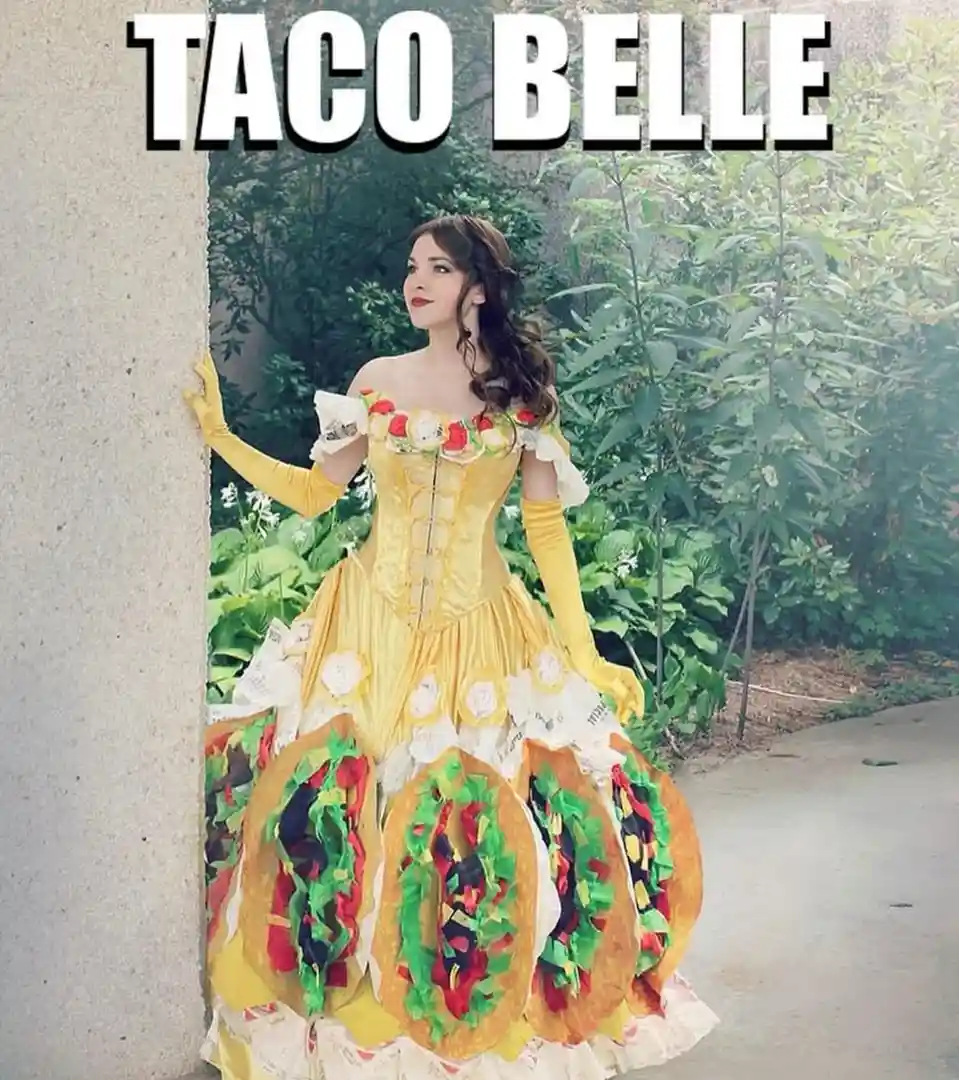 A woman in a yellow Belle-style princess dress, made to look like a taco, stands leaning against a wall outdoors. The skirt…