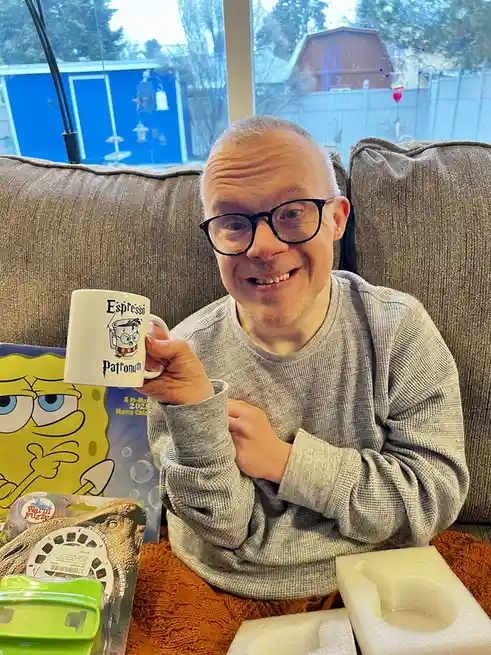 A man with glasses sits on a couch holding up a white mug that reads 'Espresso Patronum' with a cartoon drawing of a mug…