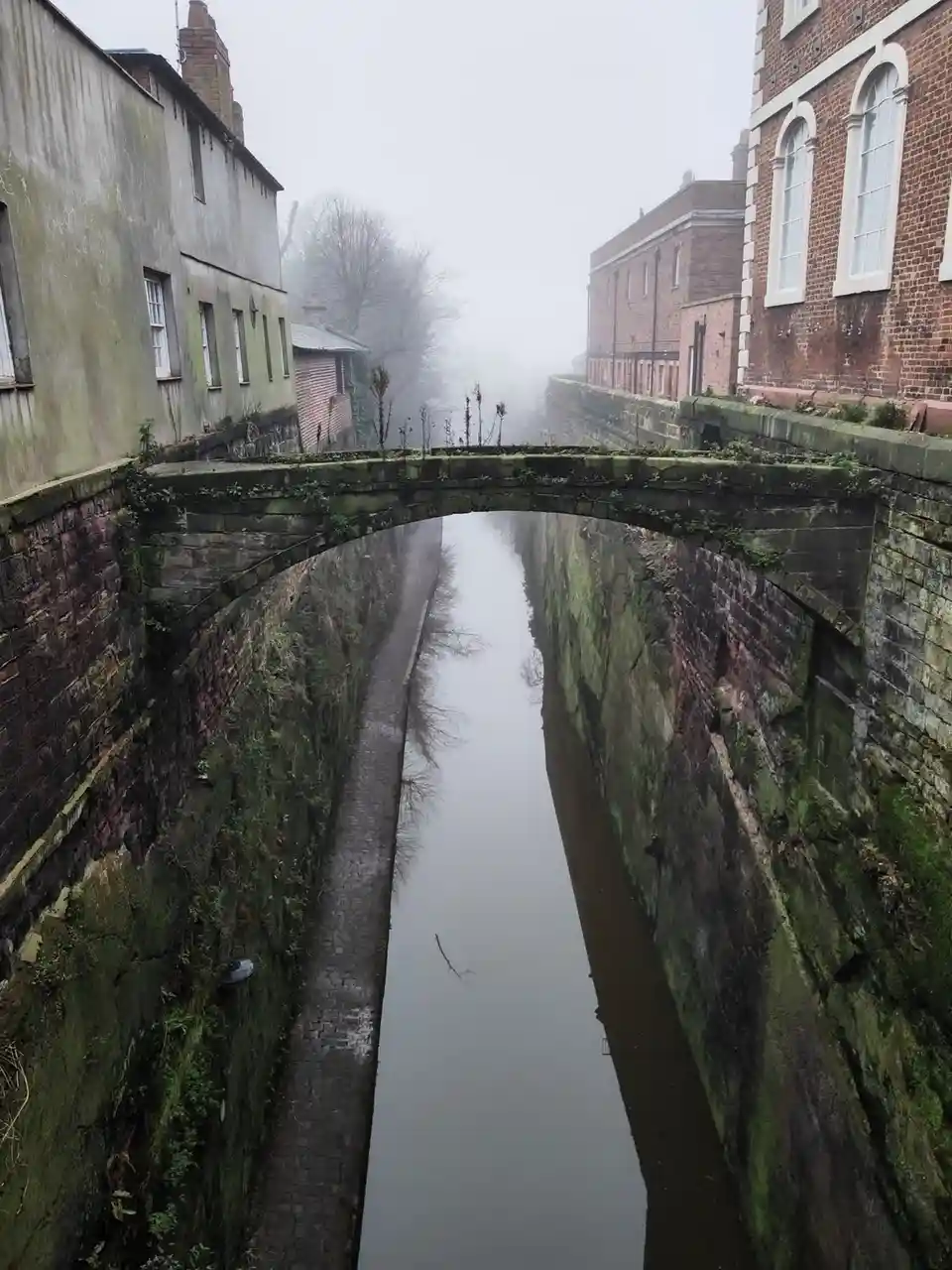 A misty, atmospheric photo of a narrow canal with a stone bridge arching over it, flanked by old brick and weathered…