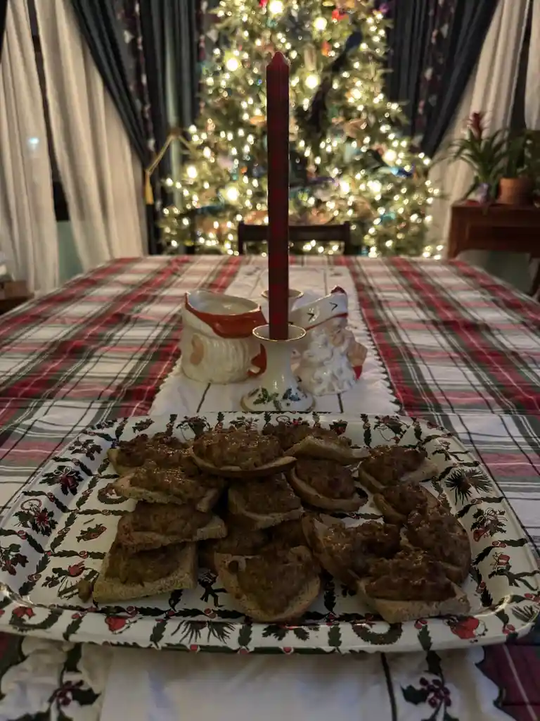 A festive Christmas table setting features a platter of bread slices topped with what appears to be meatloaf or sausage…