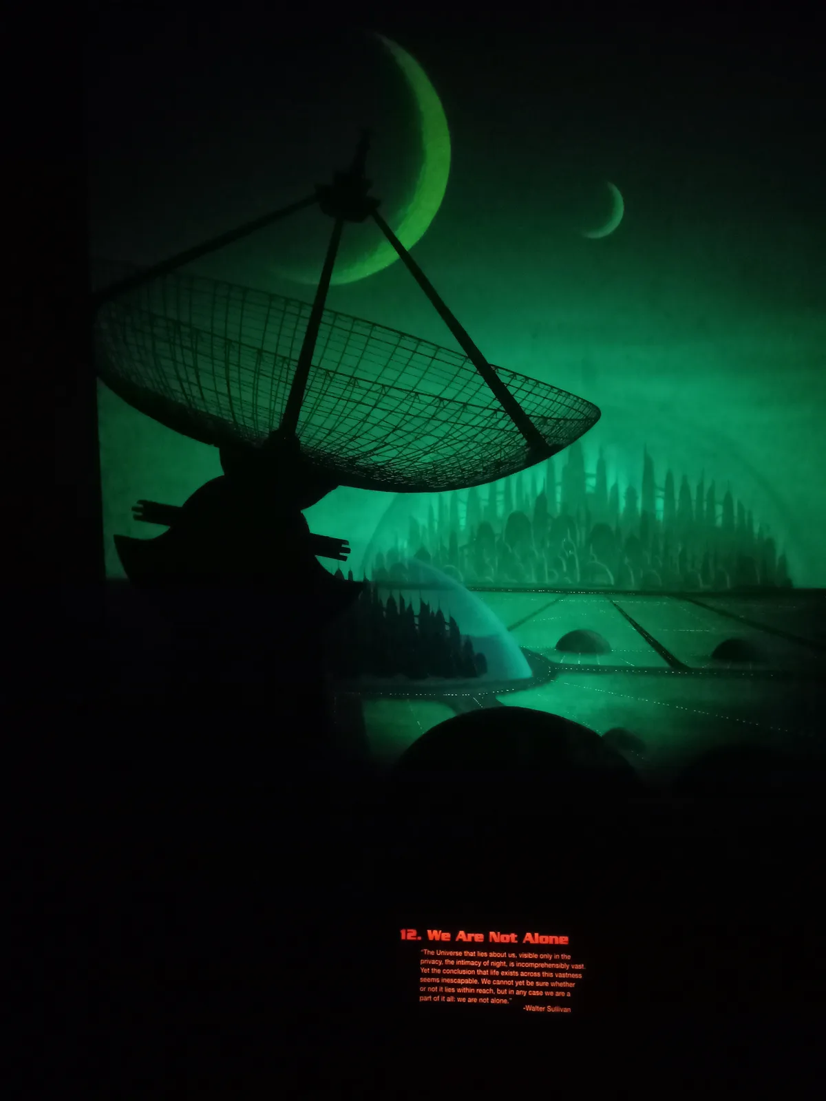 A dark, green-tinted image shows a large satellite dish silhouetted against a futuristic alien cityscape under crescent…