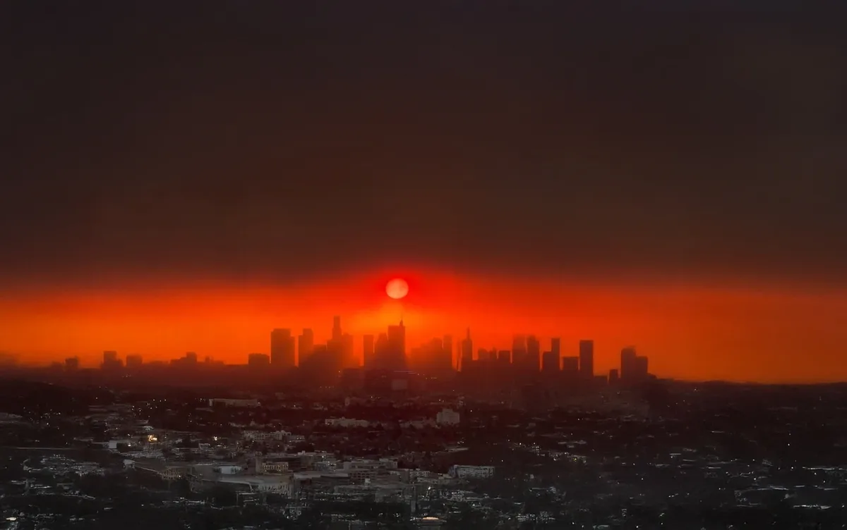 A dramatic sunset over the Los Angeles skyline, with the sun appearing as a bright orange orb just above the buildings…