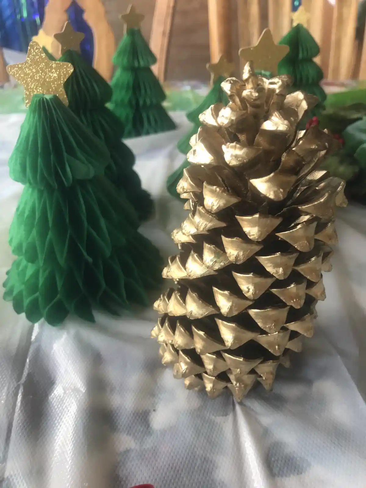 A close-up photo shows a gold-painted pinecone standing upright on a white surface, surrounded by several green paper…