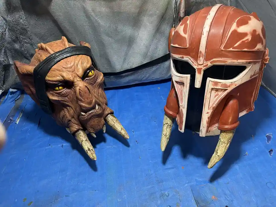 Two custom-made cosplay helmets sit side-by-side on a blue tarp: one is a detailed Mandalorian helmet with orange and white…