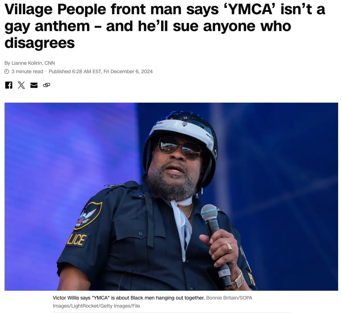 A man wearing a police-style uniform and a helmet with sunglasses holds a microphone, appearing to speak. The overlaid text…