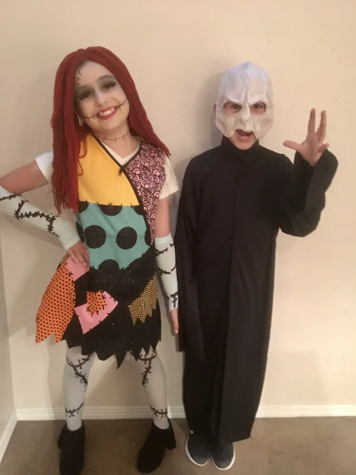 Two children dressed as Halloween characters stand against a plain wall. The child on the left wears a colorful patchwork…