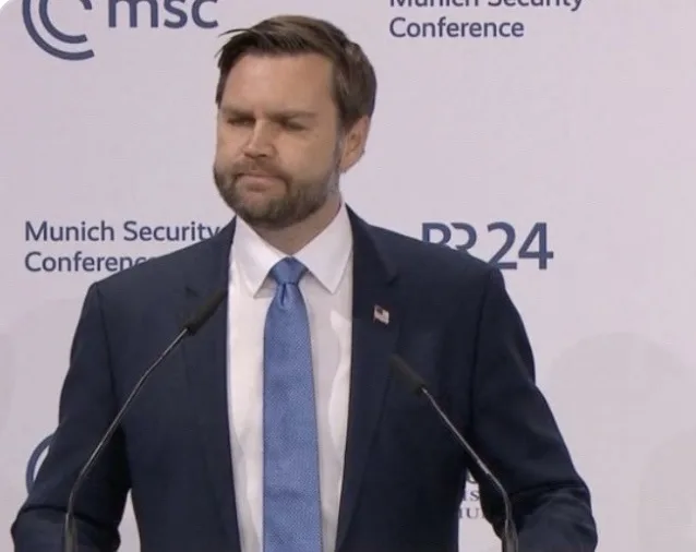 A man in a dark suit and blue tie stands at a podium with two microphones, looking down with a skeptical or unimpressed…