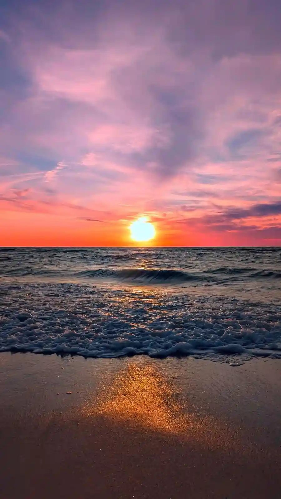 A serene beach sunset with vibrant pink, orange, and purple clouds above a calm ocean, where gentle waves wash onto wet sand…
