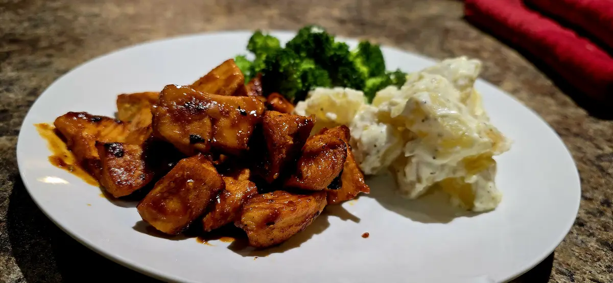 A white plate holds a meal of glazed chicken pieces, steamed broccoli, and creamy potato salad, set on a speckled countertop…
