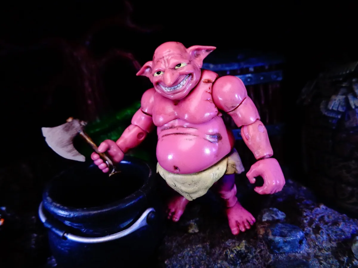 A pink, muscular, goblin-like action figure with pointed ears and a wide, toothy grin holds an axe over a black cauldron…