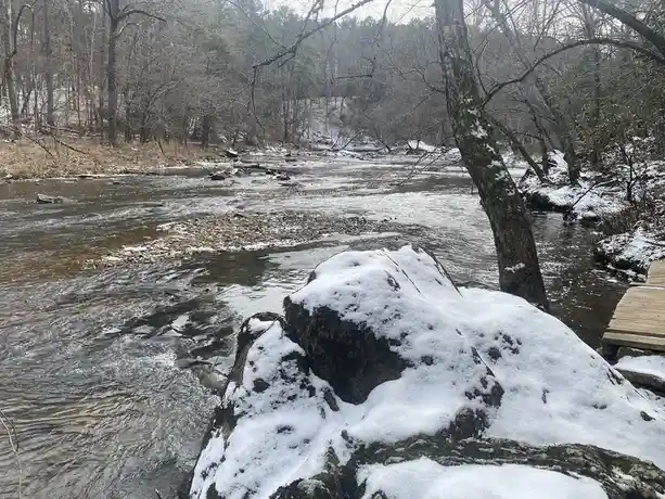 A snowy, wooded river scene with a large snow-covered rock in the foreground and a wooden boardwalk on the right. The…