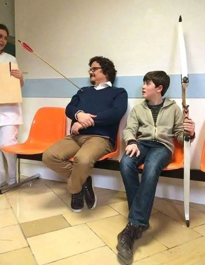 In a hospital waiting room, a man with glasses sits calmly on an orange bench with an arrow stuck in his head, while a young…