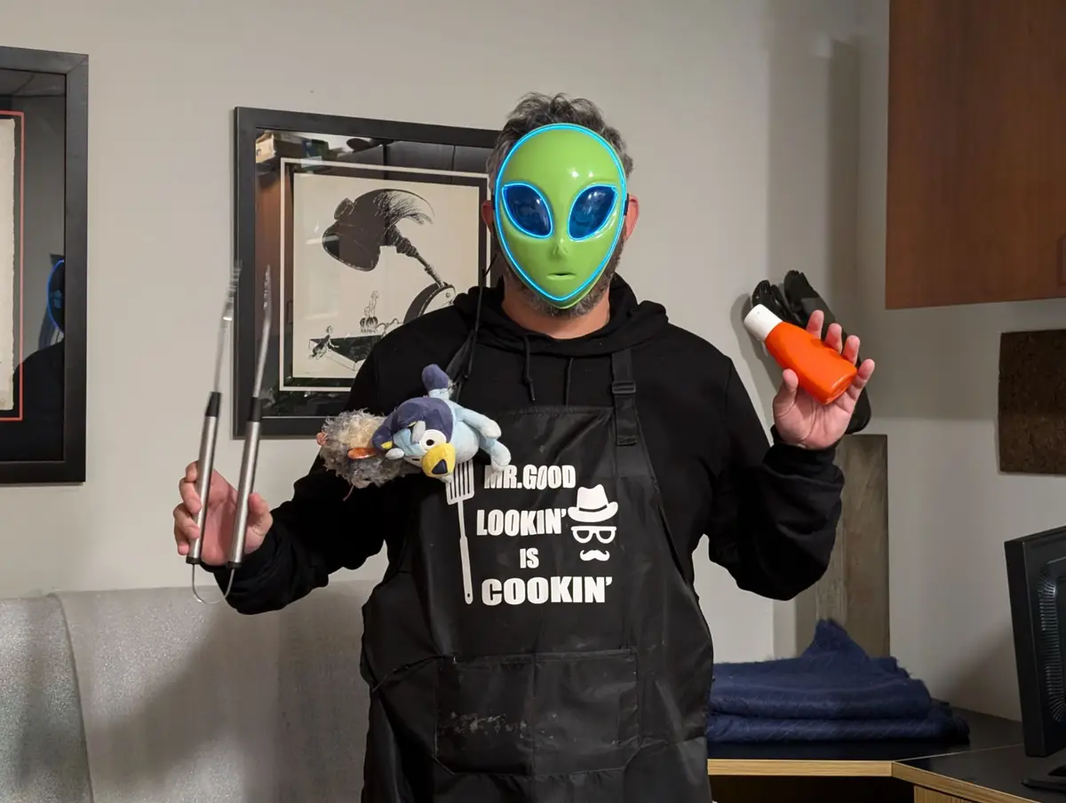 A person wearing a green alien mask with glowing blue eyes, a black apron with the text 'MR. GOOD LOOKIN' IS COOKIN'', and a…