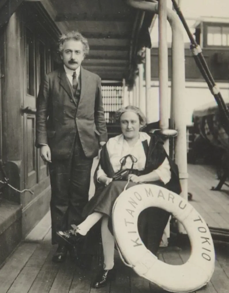 A vintage black-and-white photo shows a man and woman posing on a ship's deck. The woman sits beside a large white life…