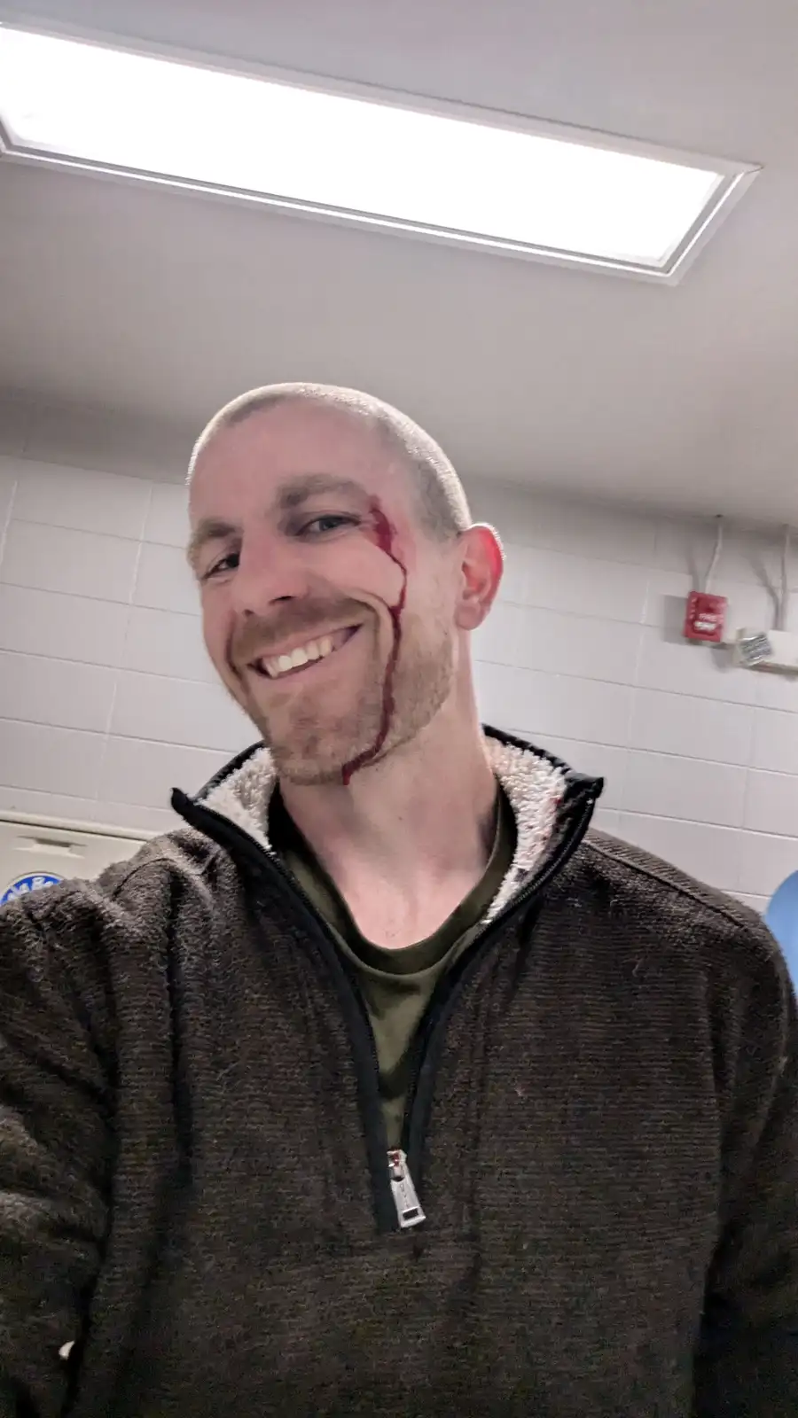 A bald man with a beard smiles widely at the camera while a streak of red paint or fake blood runs from his temple down his…