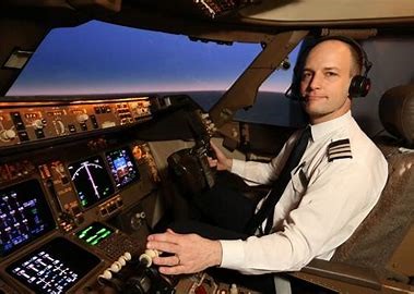 A smiling male pilot in a white uniform and headset sits in the cockpit of an airplane, hands on controls, with a sunset…