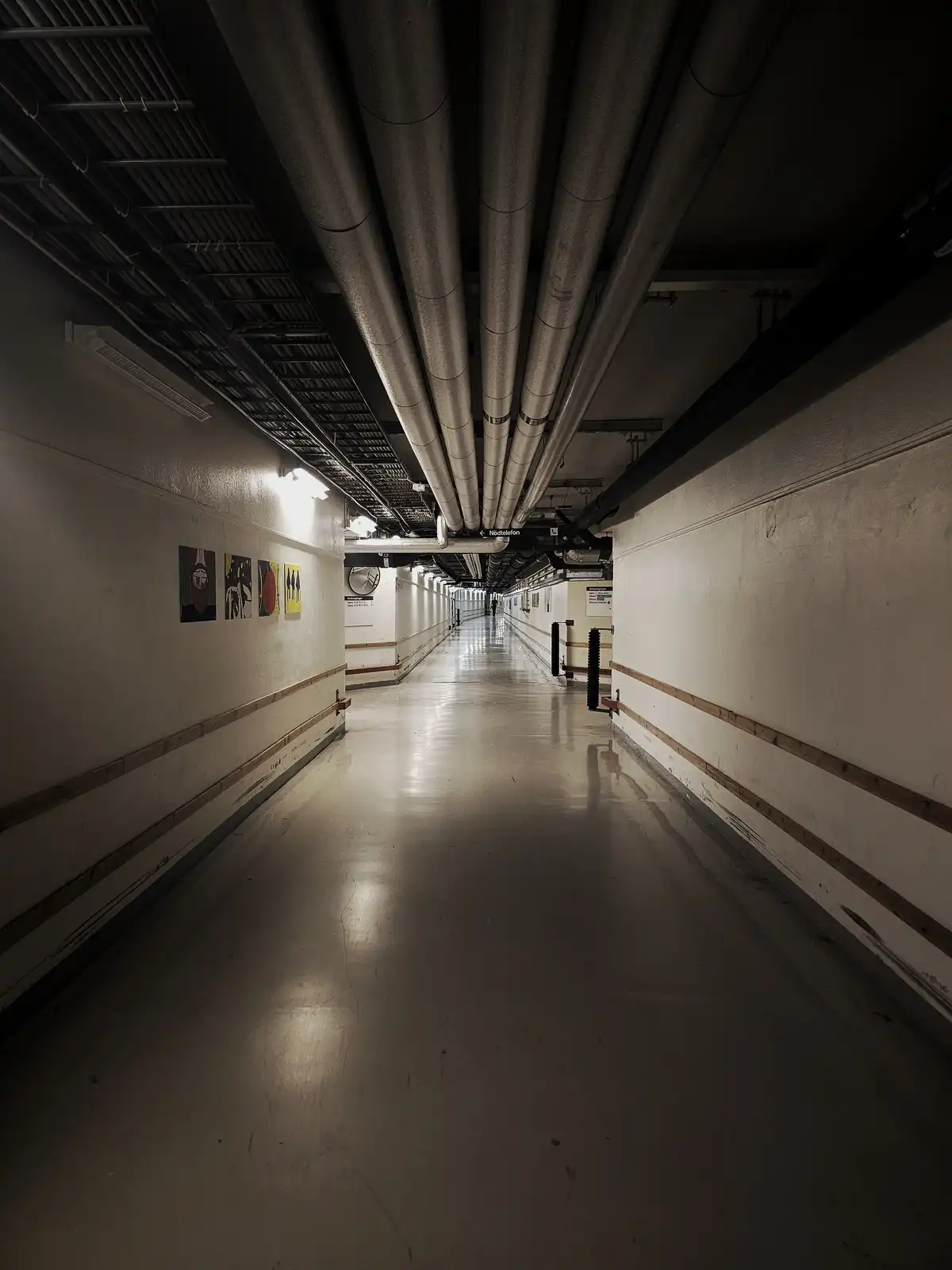 A long, dimly lit, industrial-style hallway with exposed pipes overhead and artwork on the walls, featuring a sign pointing…