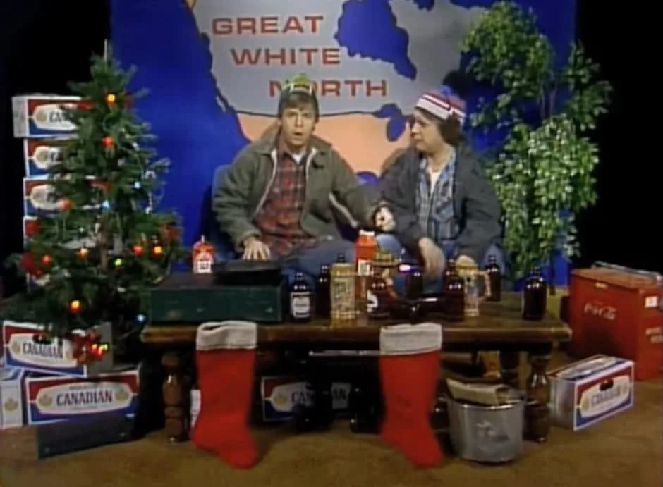 Two men sit at a cluttered table surrounded by Canadian-themed items including a decorated Christmas tree, Canadian beer…
