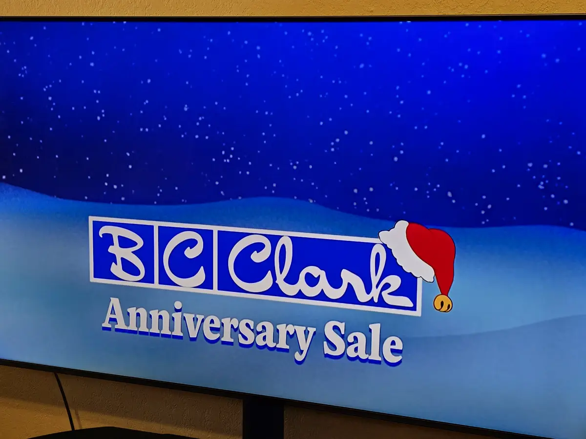 A TV screen displays a festive advertisement for 'BC Clark Anniversary Sale' against a snowy blue background. The 'BC Clark'…