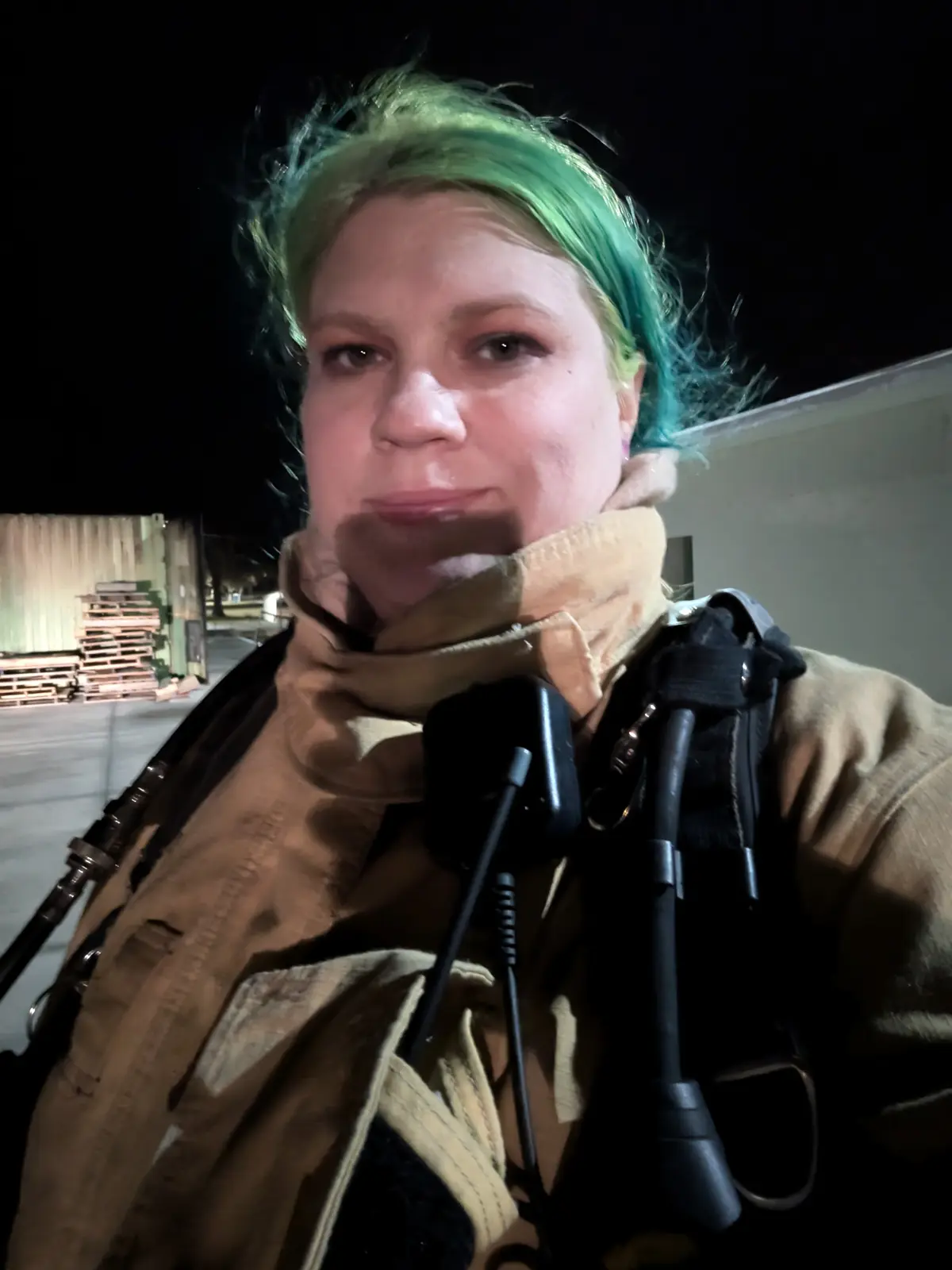 A person with bright green and blue hair, wearing a tan jacket and carrying bagpipes, looks directly at the camera with a…