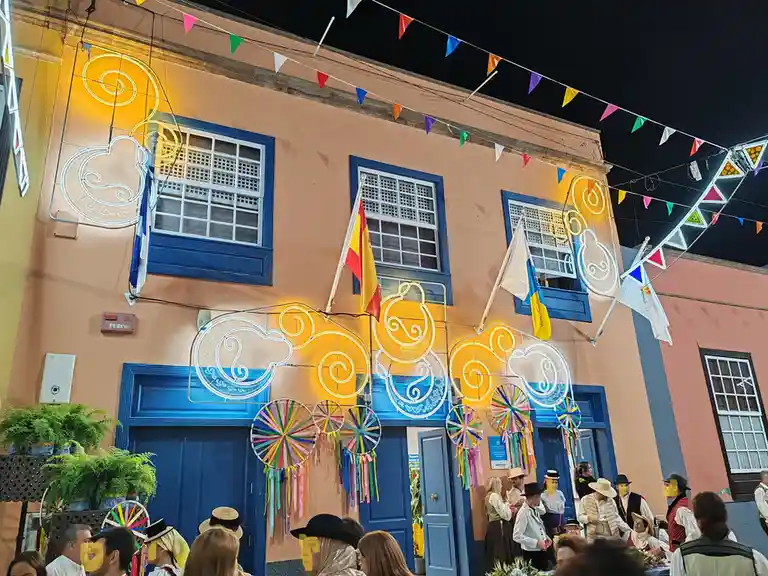 A festive night scene shows a peach-colored building with blue doors and windows, adorned with colorful string flags…