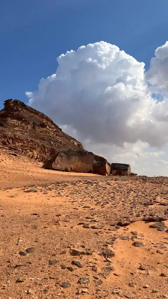 A desert landscape with orange sand and scattered rocks under a blue sky with a large, dramatic white cloud. The overlaid…