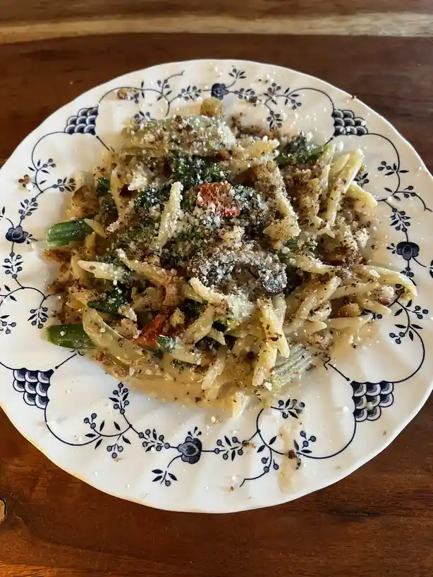 A close-up photo shows a plate of pasta with green vegetables, breadcrumbs, and grated cheese, served on a white plate with…