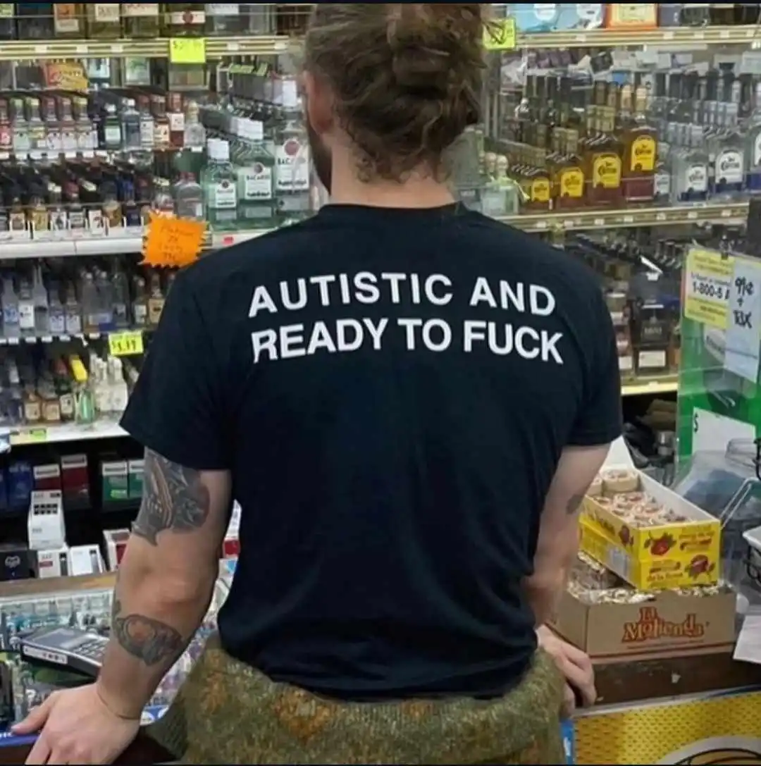 A person with long hair tied back and tattoos on their arms is seen from behind, wearing a black t-shirt with the text…