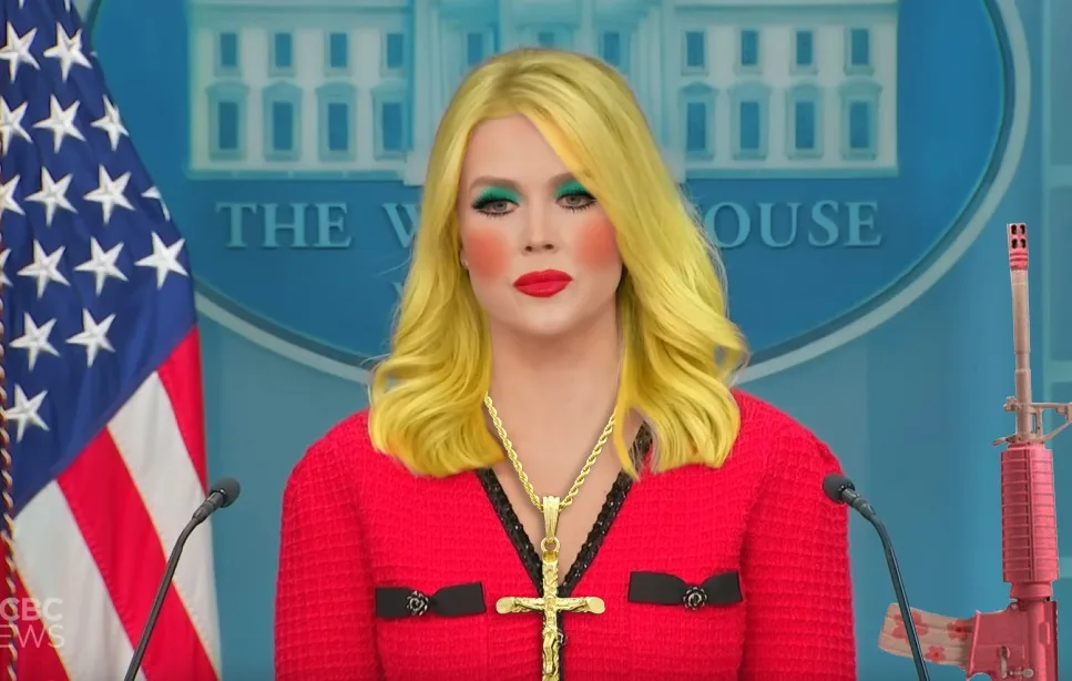a person with bright yellow hair, heavy makeup, and a red outfit stands at a podium in front of a blue backdrop with 'THE…