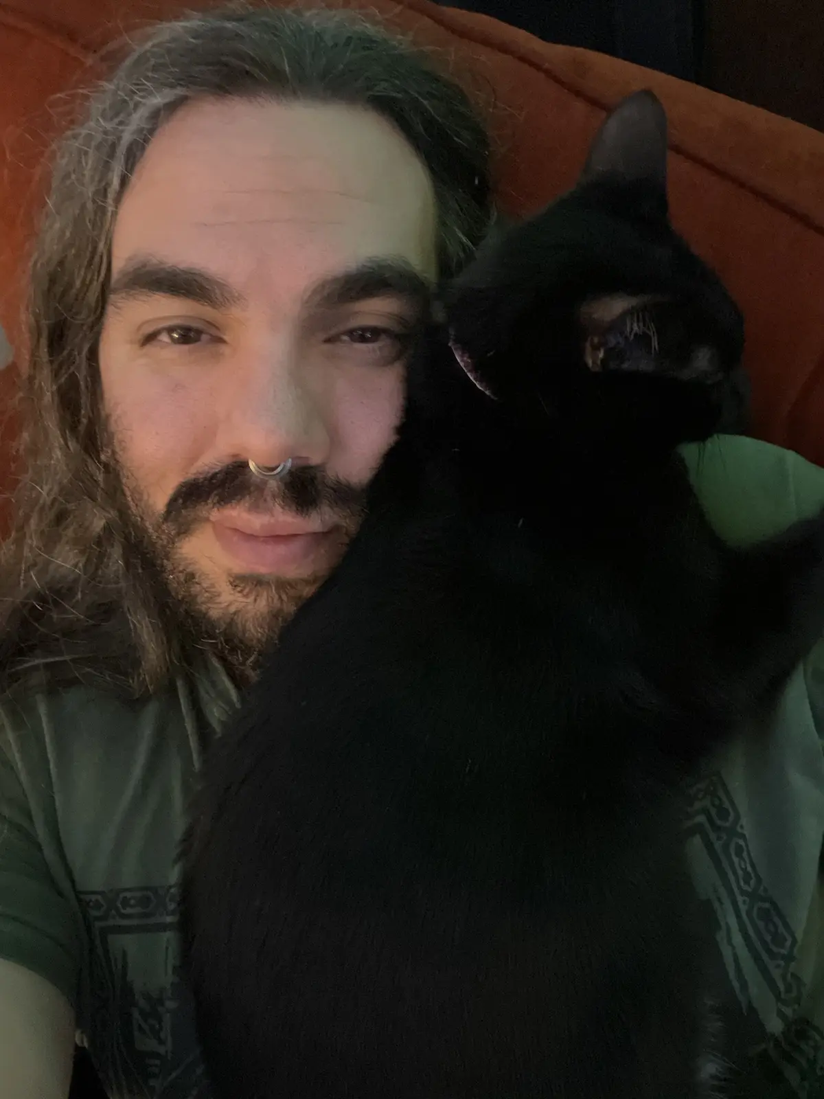 A man with long hair, a nose ring, and a beard is lying on an orange couch, smiling at the camera while holding a black cat…