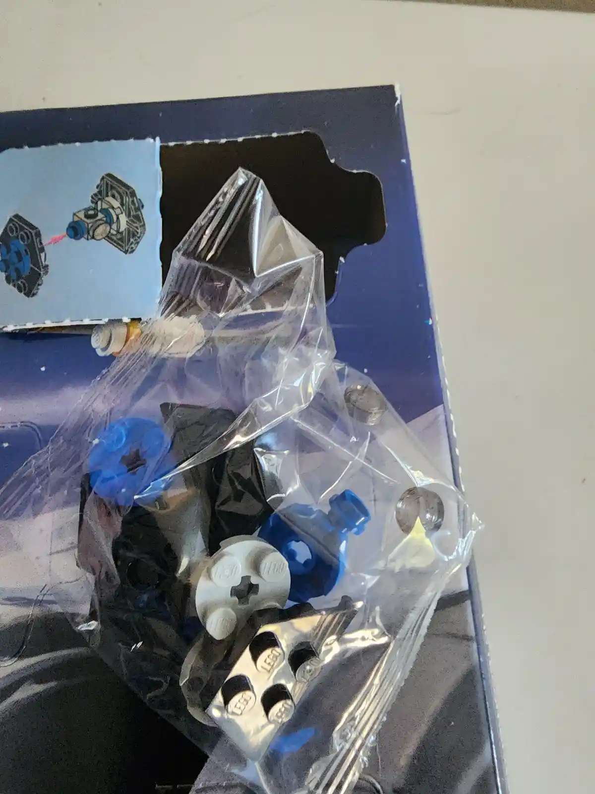 A close-up photo shows a partially opened LEGO set with pieces spilling out of a torn plastic bag onto the box's printed…