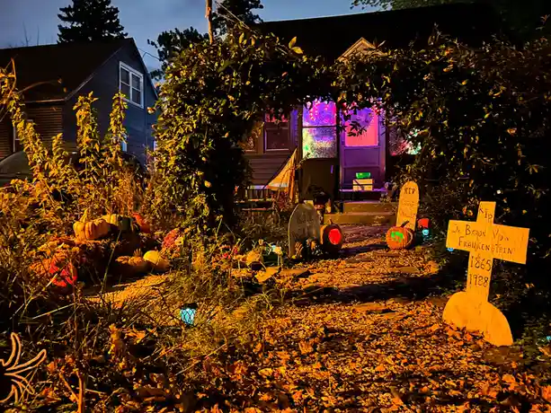 A nighttime photo of a Halloween-decorated front yard with fake tombstones, pumpkins, and glowing lights. One prominent…