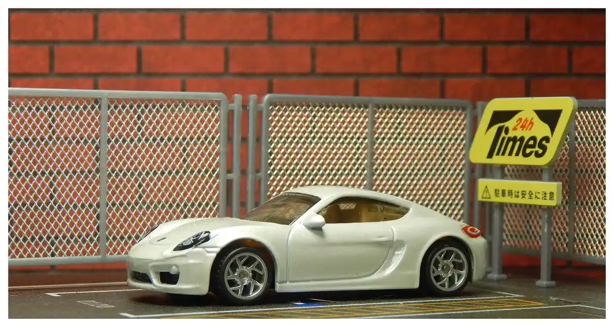 A silver toy Porsche Cayman model car is parked in a miniature parking lot with white and yellow lines, in front of a grey…